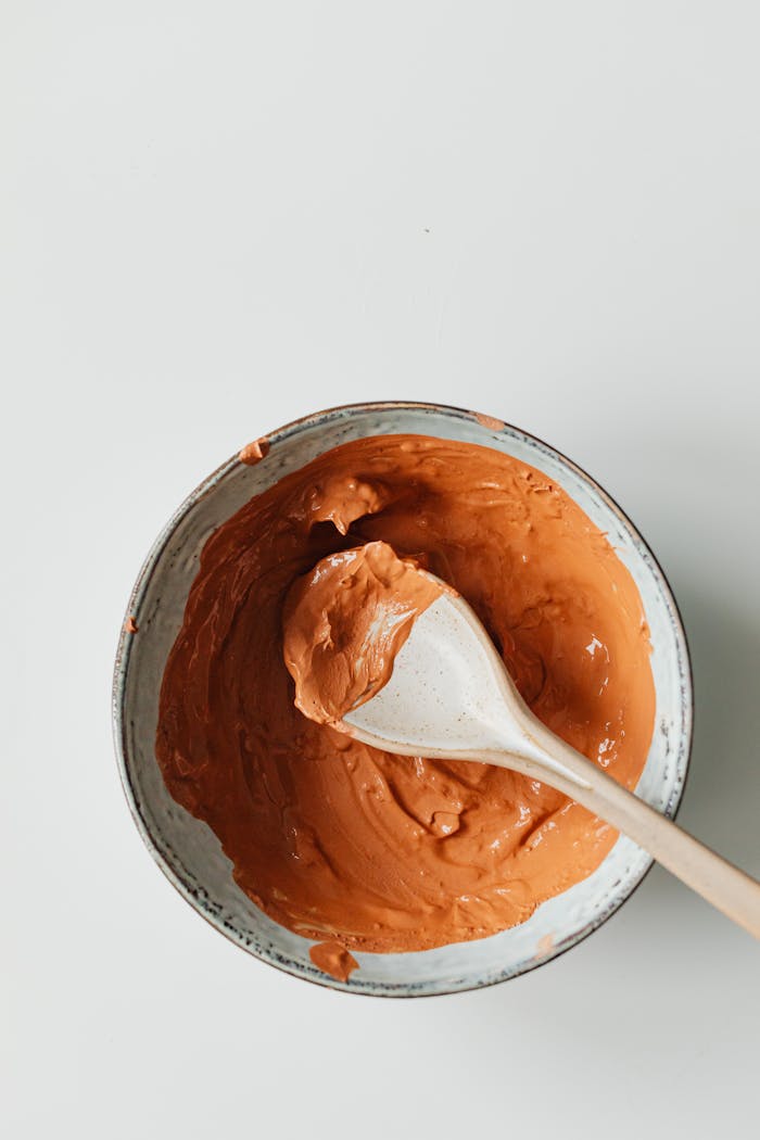 Overhead view of orange clay mask in a ceramic bowl with a wooden spoon, ideal for skincare.